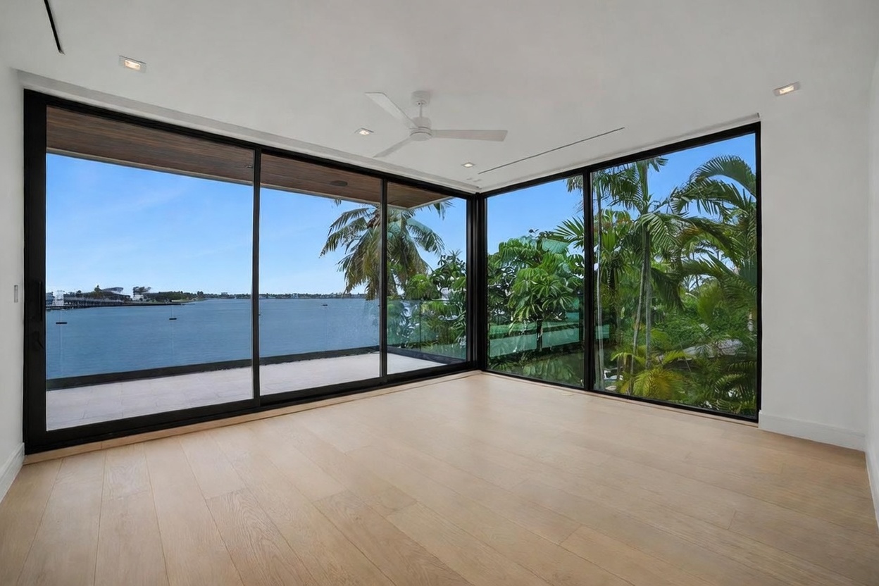 This bright, minimalist room features expansive floor-to-ceiling glass walls that provide an unobstructed, panoramic view of the water and lush tropical foliage. The space is finished with light wood flooring and a clean, white ceiling equipped with a modern ceiling fan. The perspective captures the seamless transition between the interior living space and the outdoor environment, emphasizing a serene and luxurious atmosphere.