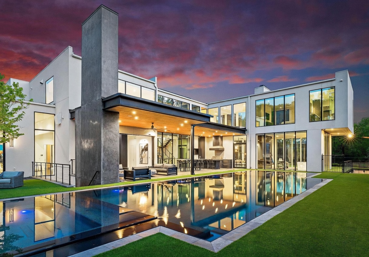 This modern luxury residence features a striking rear facade with clean lines and extensive glass walls that connect the indoor and outdoor living spaces. A large, covered patio area with a wood-paneled ceiling and an outdoor kitchen overlooks a sleek, rectangular infinity-edge pool. The scene is set against a dramatic sunset sky, highlighting the architectural contrast between the dark, tall chimney structure and the light-toned exterior walls.