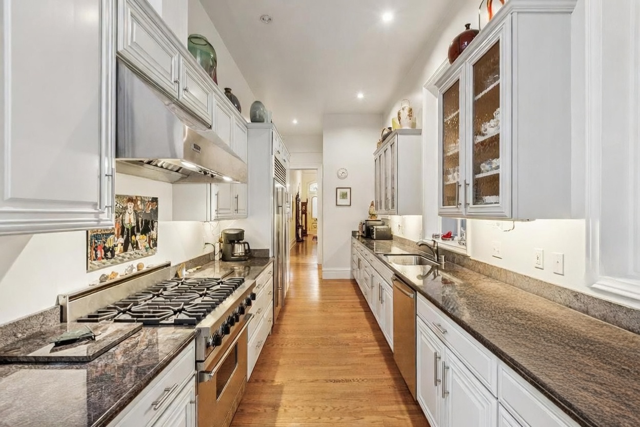 This galley-style kitchen features white cabinetry paired with dark, patterned stone countertops and stainless steel appliances, including a professional-grade range. The space is illuminated by under-cabinet lighting and recessed ceiling lights, highlighting the warm wood flooring that runs throughout. A long, narrow layout leads the eye toward a doorway at the far end, creating a clean and functional culinary workspace.