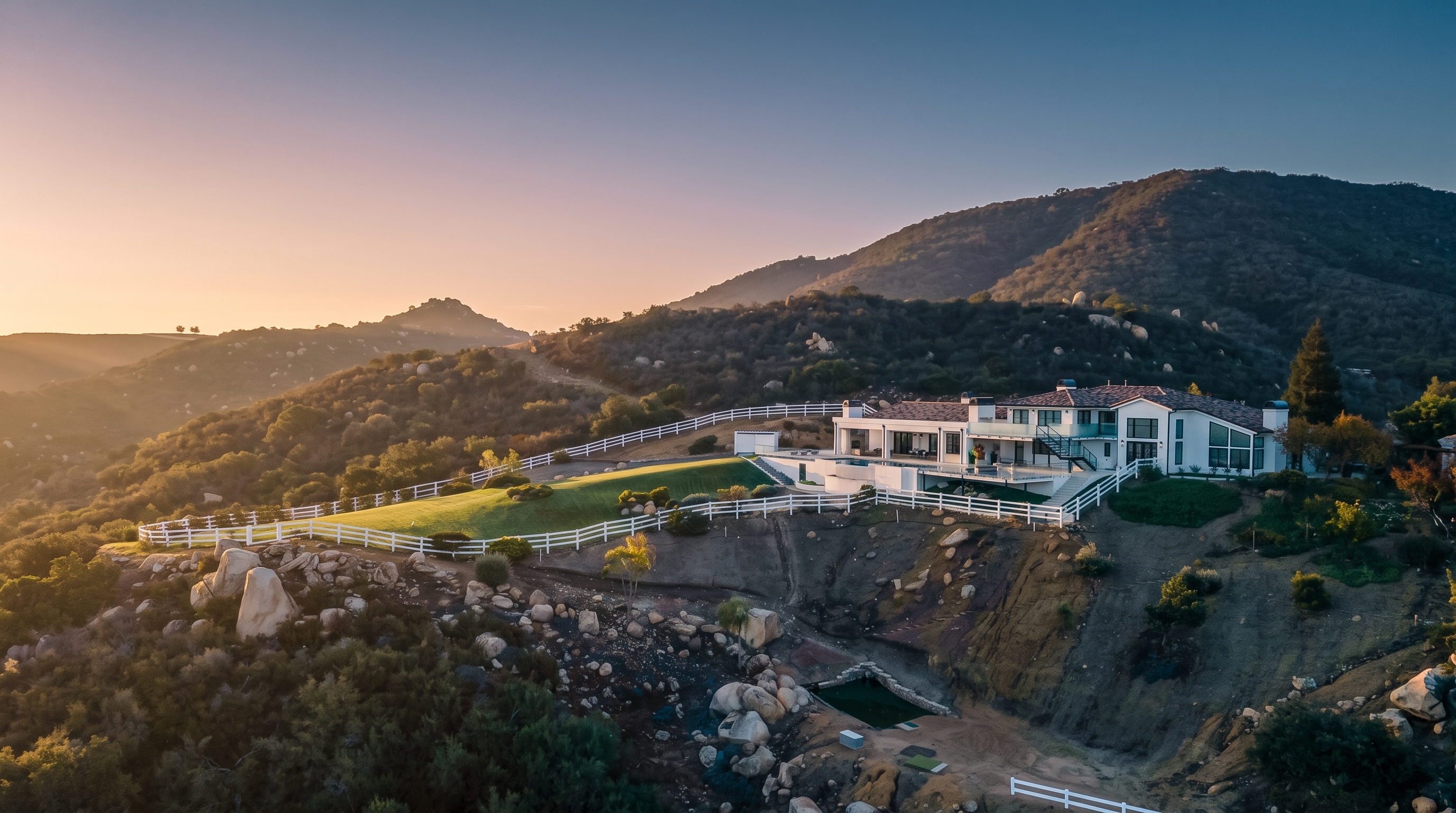 This stunning aerial view showcases a luxurious, sprawling modern estate perched prominently on a hillside, overlooking vast, rugged terrain at sunset. The property features an expansive lawn enclosed by white fencing, a multi-level terrace, and high-end architectural design with large windows and a terracotta-style roof. The dramatic landscape and vibrant pink and purple twilight sky create a sophisticated, exclusive atmosphere perfect for a high-end real estate listing.