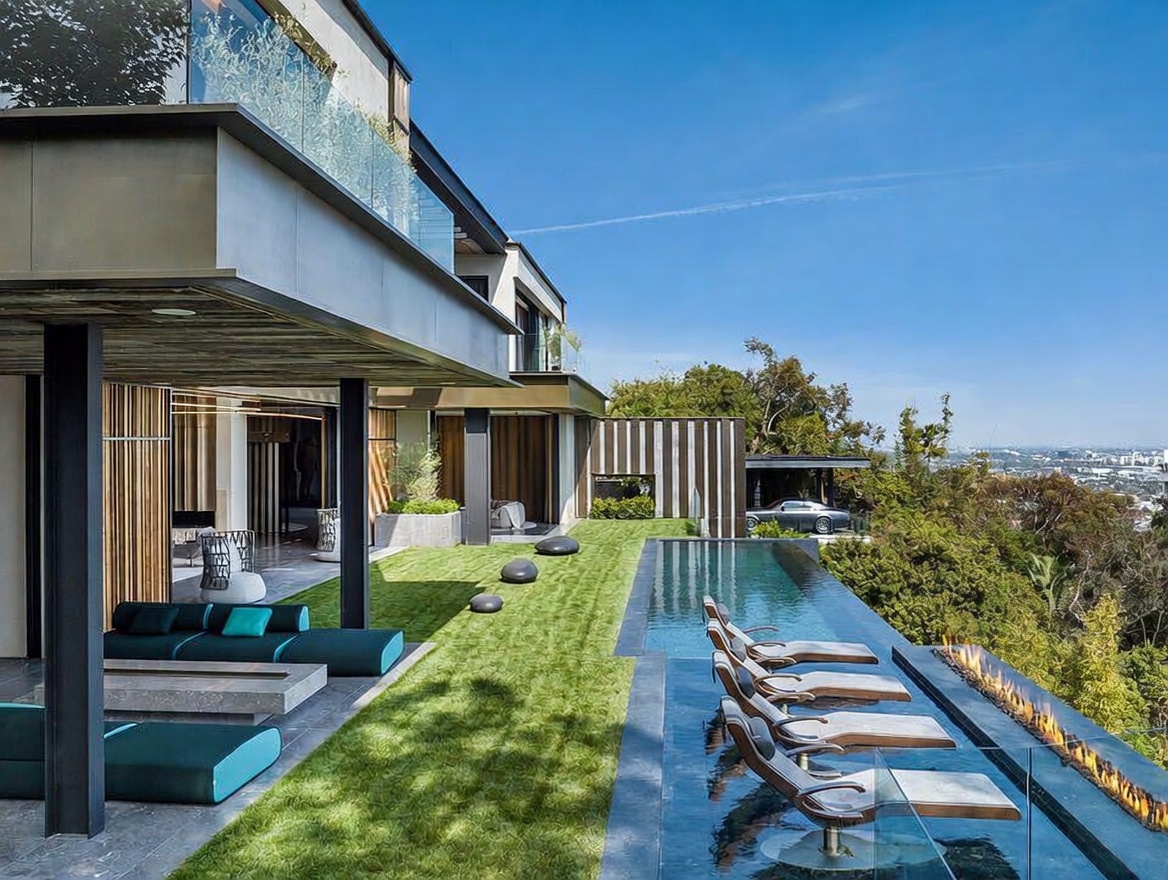 This stunning modern luxury estate features a seamless indoor-outdoor living space with a sprawling lawn and a sleek infinity-edge pool overlooking a panoramic city view. The patio area is furnished with contemporary lounge seating and a fire feature, creating an ideal environment for high-end entertaining. The architectural design emphasizes clean lines, expansive glass, and a sophisticated, minimalist aesthetic.