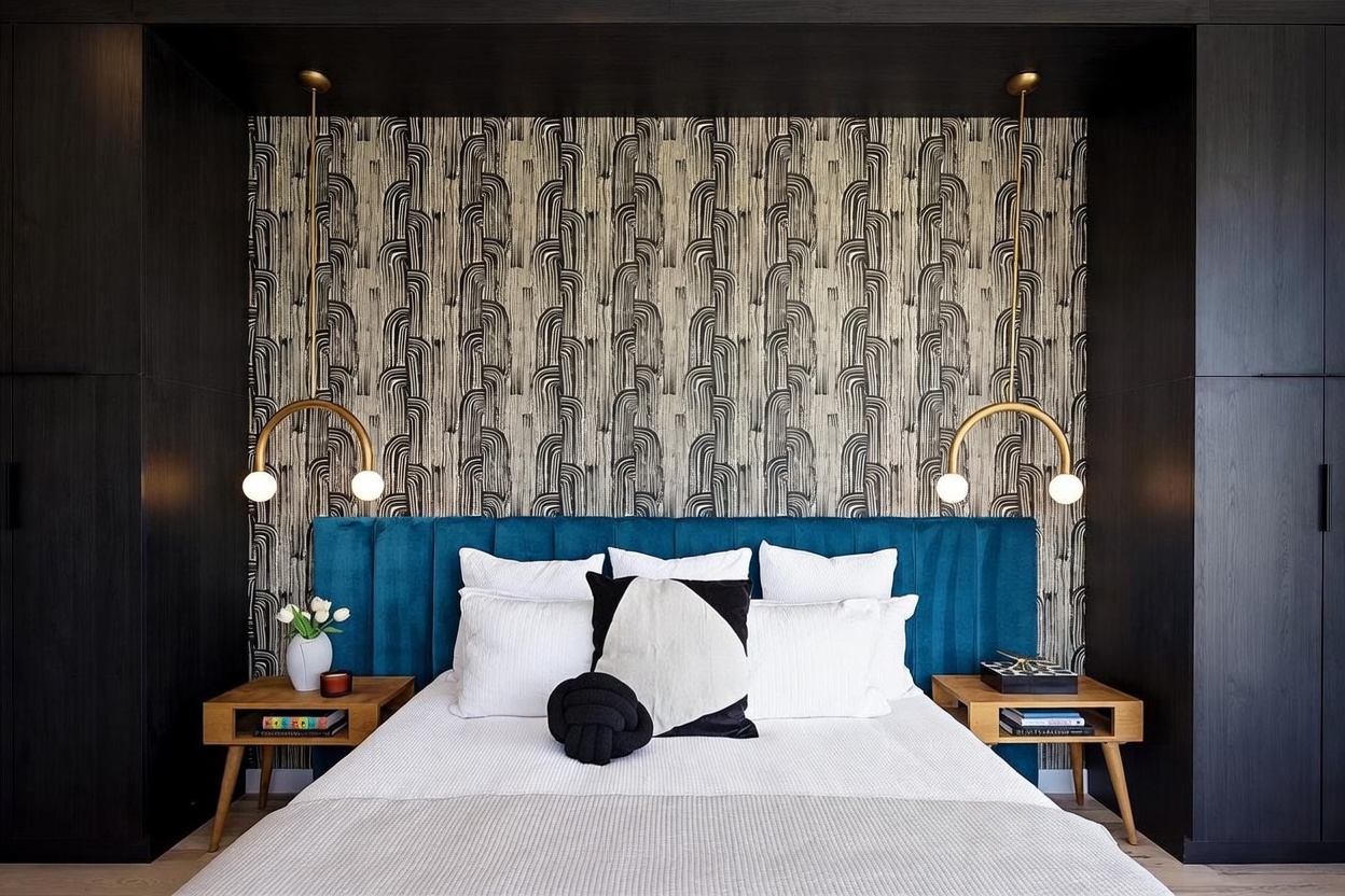 This primary bedroom features a bold, modern aesthetic centered around a plush, deep blue velvet headboard set against a striking, patterned wallpaper. The bed is flanked by two light wood nightstands and unique, arched gold pendant lights that provide a warm, focused glow. The room is framed by dark, built-in cabinetry on either side, creating a sophisticated and symmetrical focal point.