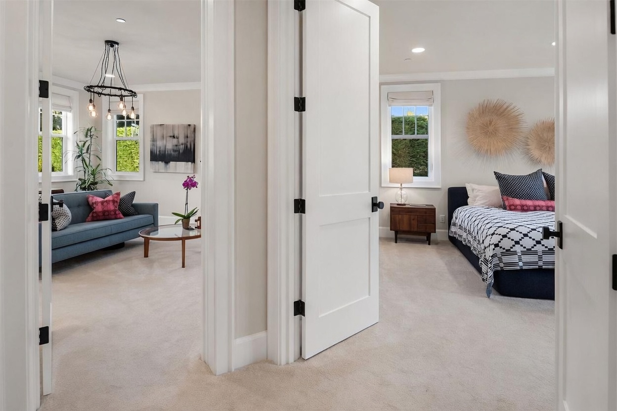 This perspective captures a transition space between two rooms, featuring a white door frame that separates a living area on the left from a bedroom on the right. The living room is furnished with a blue sofa, a glass-topped coffee table, and a decorative chandelier, while the bedroom showcases a bed with patterned bedding and wall-mounted circular art. The neutral color palette and consistent carpeting create a cohesive and inviting flow throughout the home.