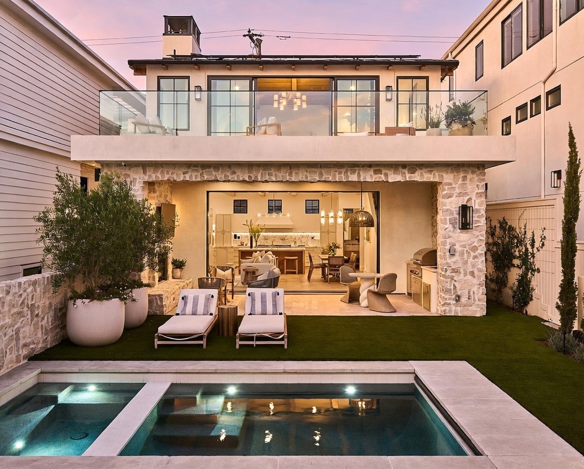 This luxurious rear exterior view showcases a modern home with a seamless indoor-outdoor transition, featuring a large open-concept living space that flows onto a stone-paved patio. The backyard includes a pristine rectangular swimming pool, a manicured lawn, and a stone-clad wall that adds texture to the contemporary design. Above, a spacious balcony with a glass railing provides an elevated vantage point, while the warm interior lighting creates an inviting atmosphere at dusk.