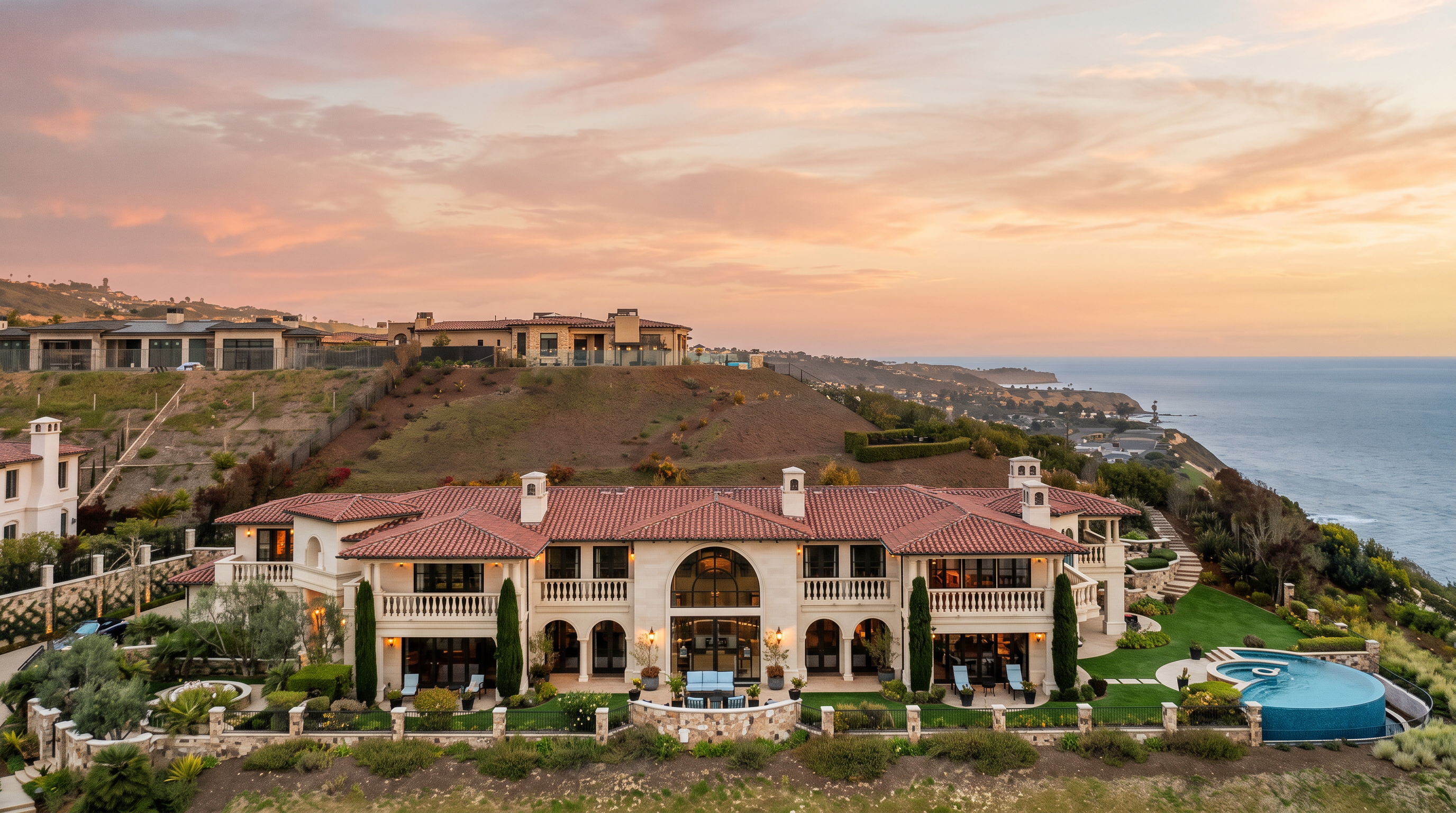 This stunning aerial perspective captures a sprawling Mediterranean-style estate perched on a coastal cliffside at sunset. The residence features a classic terracotta tile roof, expansive balconies, and a curved infinity-edge pool that overlooks the vast ocean horizon. The warm, vibrant colors of the twilight sky create a dramatic backdrop for this luxurious property, emphasizing its secluded and prestigious location.