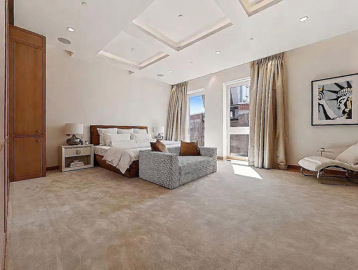 This spacious primary bedroom features a neutral color palette, plush carpeting, and a coffered ceiling with recessed lighting. A large bed with a wooden headboard is positioned centrally, complemented by a patterned bench at the foot and a modern lounge chair near the window. Two tall windows with floor-to-ceiling curtains allow for ample natural light, creating an airy and sophisticated atmosphere.