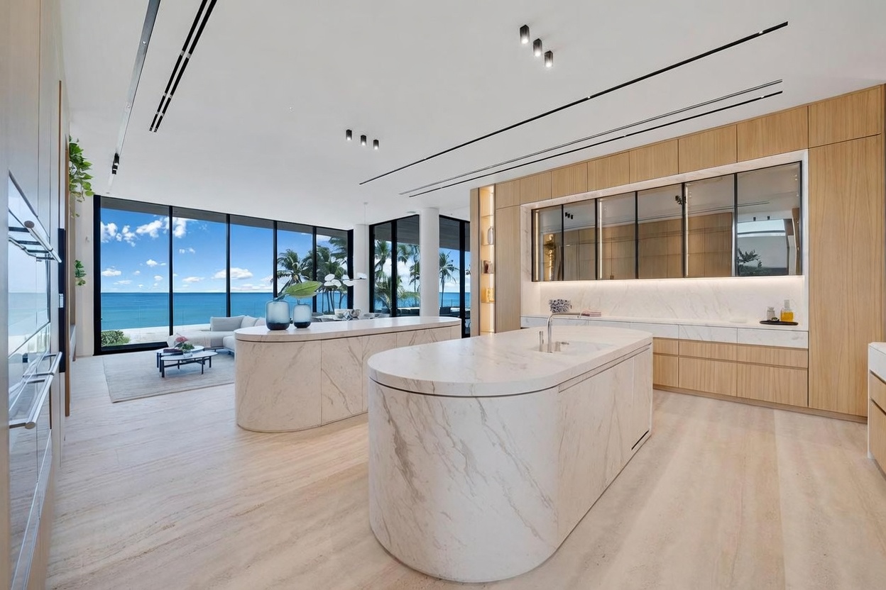 This expansive, modern kitchen features two large, curved islands with light-toned stone surfaces and integrated cabinetry. The space is defined by light wood cabinetry, sleek reflective panels, and floor-to-ceiling windows that provide an unobstructed view of the ocean. The open-concept design seamlessly transitions into a living area, creating a bright, airy atmosphere with a minimalist aesthetic.