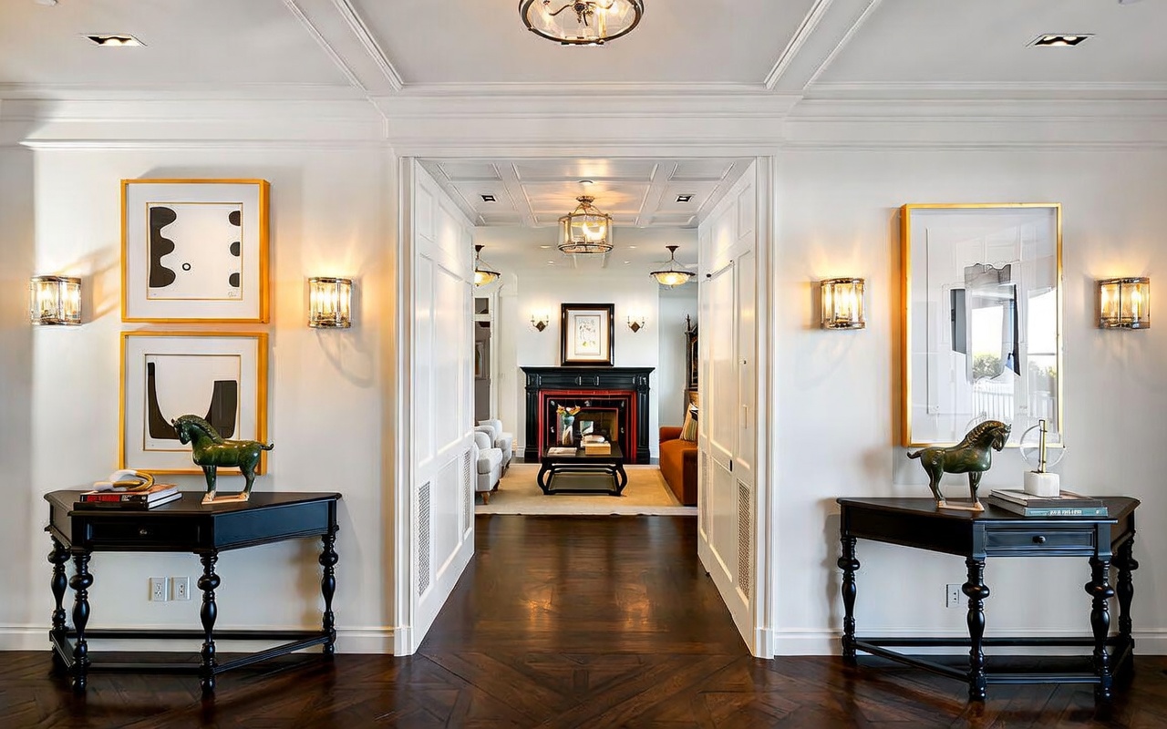 This elegant foyer features a symmetrical layout with dark wood flooring arranged in a herringbone pattern, leading the eye toward a distant living area with a fireplace. The space is framed by two matching black console tables topped with horse sculptures and framed artwork, flanked by wall-mounted sconces. The bright, white-walled hallway creates a sophisticated transition between rooms, enhanced by recessed lighting and a central ceiling fixture.