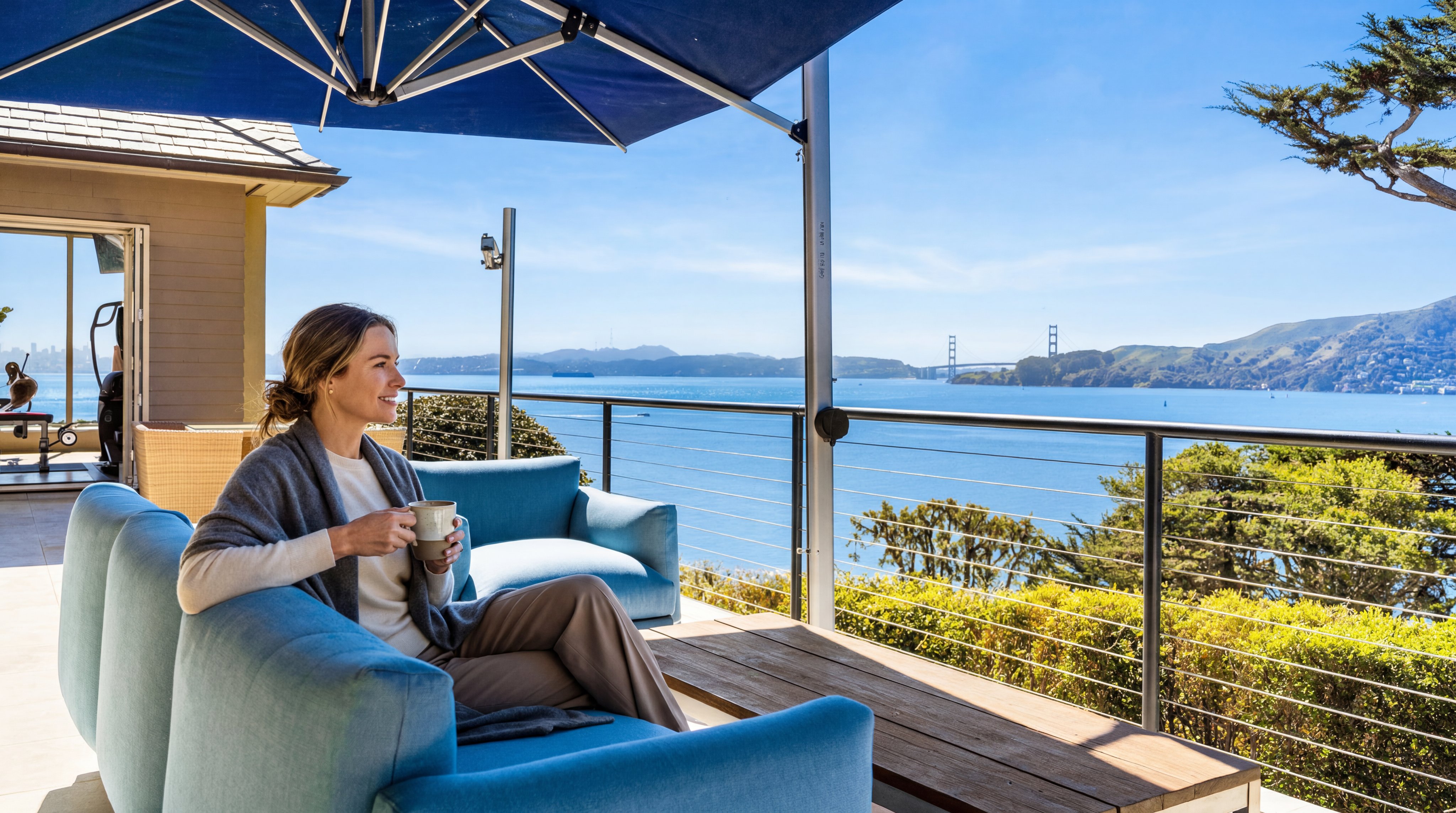 This expansive outdoor terrace offers a breathtaking, unobstructed view of the Golden Gate Bridge and the surrounding bay. The space is furnished with comfortable blue lounge seating and a wooden coffee table, all shaded by a large, modern blue umbrella. The perspective captures the seamless transition between the indoor living area and the scenic deck, emphasizing the property's prime waterfront location.