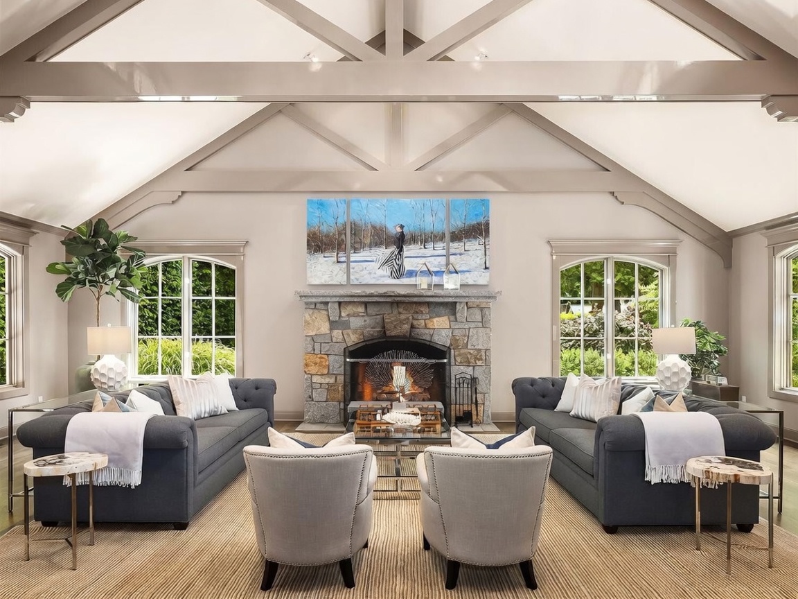 This elegant living room features a symmetrical layout with two dark gray sofas facing each other, centered around a stone-clad chimney breast and a central fireplace. The room is defined by its dramatic vaulted ceiling with exposed structural beams and large, arched windows that flood the space with natural light. A neutral color palette, textured area rug, and two light-toned armchairs create a sophisticated and inviting atmosphere.
