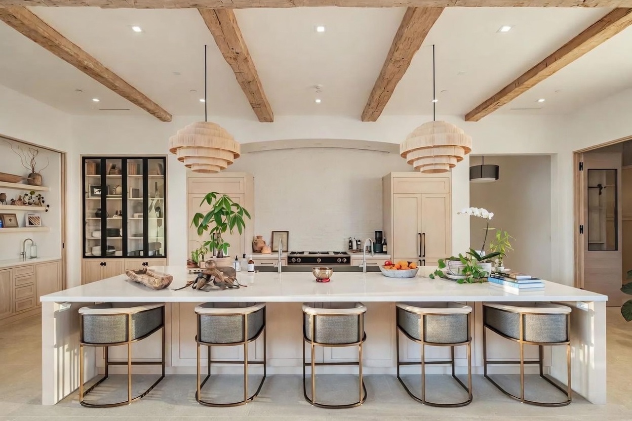 This sophisticated kitchen features a massive central island with a light-toned stone countertop and five modern bar stools with metallic frames. The space is defined by exposed wooden ceiling beams, a neutral color palette, and a symmetrical layout that includes a built-in range set against a textured white wall. To the left, a glass-fronted display cabinet adds a touch of elegance, while two large, tiered pendant lights anchor the island area, creating a warm and inviting atmosphere.