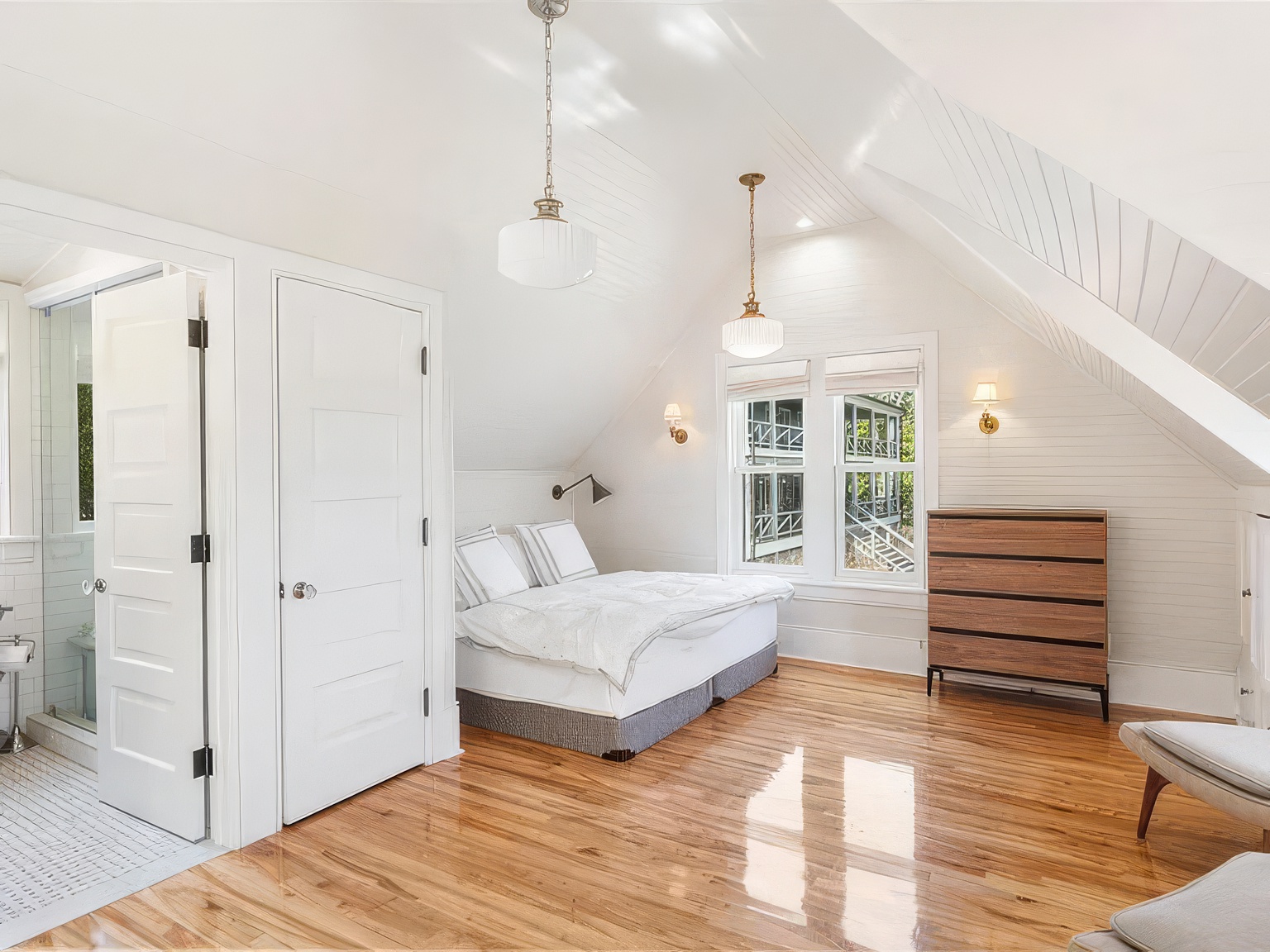 This bright, airy bedroom features a vaulted ceiling with white shiplap paneling and polished light wood flooring. The room is furnished with a low-profile bed, a dark wood dresser, and two modern pendant lights, while a doorway leads directly into an adjacent bathroom. The perspective is from the doorway, capturing the clean, coastal-inspired aesthetic and the natural light streaming through the windows.