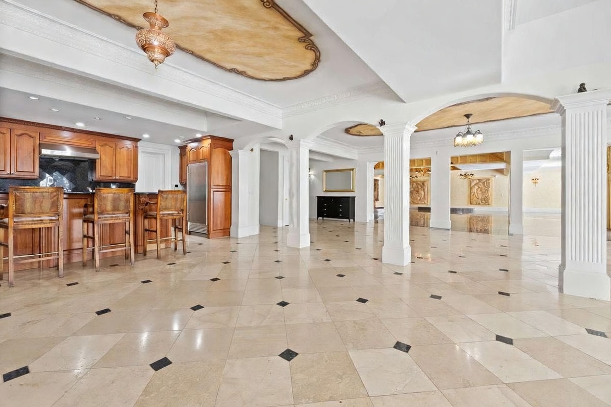 This expansive open-concept kitchen and living area features a large wooden island with bar seating and a matching cabinetry wall. The space is defined by ornate ceiling details, including recessed tray ceilings with decorative molding and hanging light fixtures, set against a backdrop of light-colored stone flooring with dark accent tiles. Multiple white columns create a grand, architectural transition between the kitchen and the adjacent living space, which includes a black console table and a distant view of a formal room.
