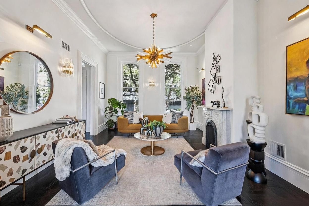 This elegant living room features dark hardwood floors, a mustard-colored sofa, and two plush grey armchairs arranged around a circular stone-topped coffee table. A prominent starburst chandelier hangs from the center of the room, while a white fireplace with a decorative mantel serves as a focal point on the right wall. The space is accented by a large round mirror, a patterned sideboard, and vibrant artwork, creating a sophisticated and well-curated atmosphere.