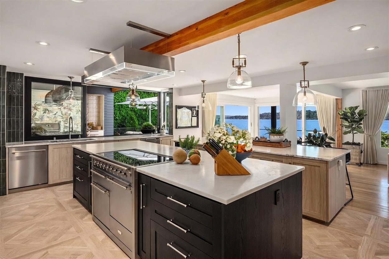 This expansive, modern kitchen features a dual-island layout with dark cabinetry and light stone countertops, centered around a professional-grade stainless steel range. A large, industrial-style vent hood hangs above the primary island, while a warm wood beam spans the ceiling, adding a rustic touch to the contemporary design. The space opens up to a scenic waterfront view through large windows, creating a bright and airy atmosphere that seamlessly blends indoor luxury with the natural landscape.