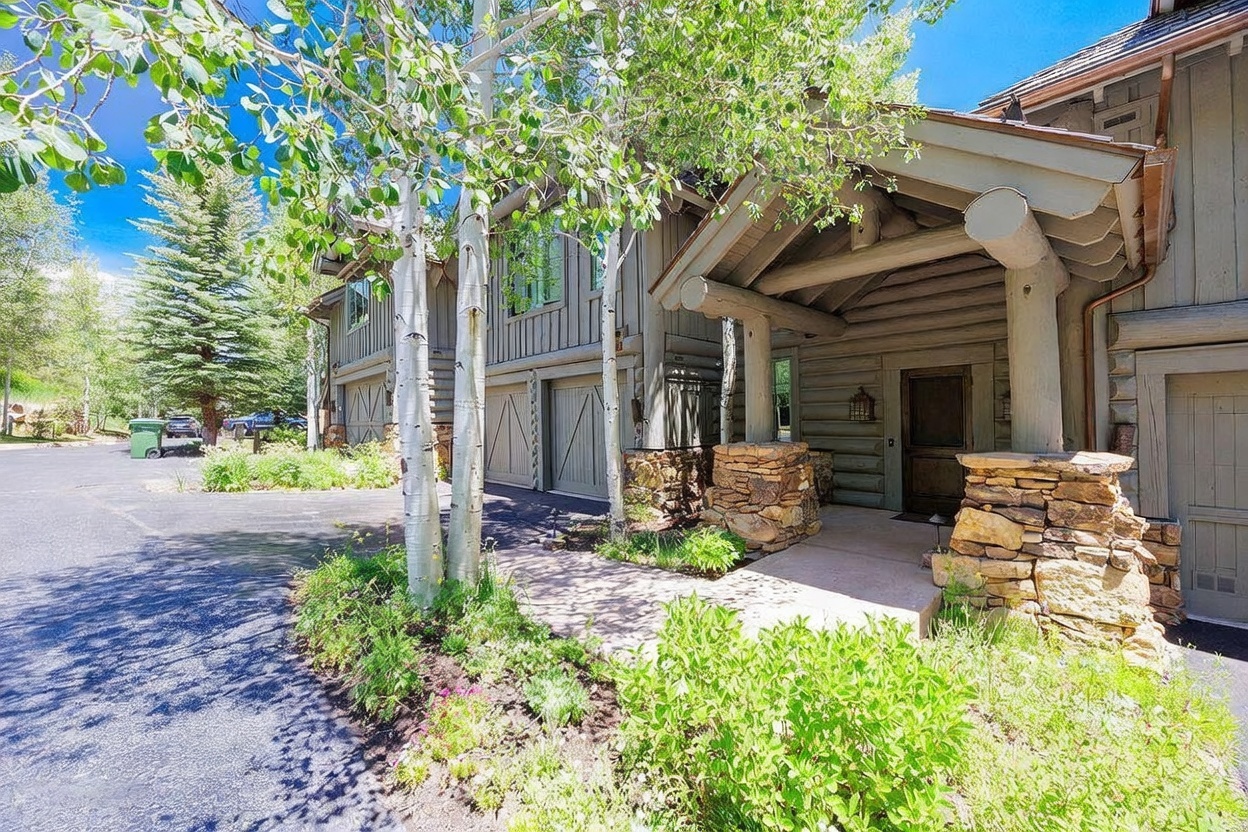 This mountain-style residence features a welcoming entryway highlighted by a covered porch supported by heavy timber posts and rustic stone pillars. The exterior is clad in vertical wood siding, complemented by a paved driveway and lush, mature landscaping including birch trees and vibrant green shrubs. The perspective captures the home's inviting curb appeal, emphasizing its natural aesthetic and integration with the surrounding wooded environment.