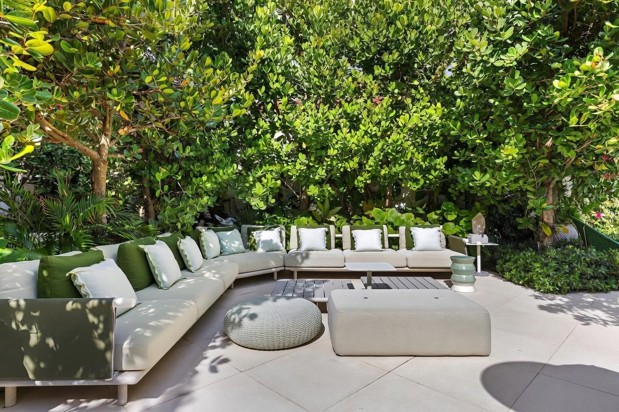 This inviting outdoor patio space features a large, L-shaped sectional sofa with light-colored cushions and green accent pillows, arranged around a low, modular coffee table. A round, textured ottoman and a square footstool provide additional seating on the paved surface, all nestled against a backdrop of lush, dense greenery. The scene is captured from a slightly elevated perspective, emphasizing the private, tranquil atmosphere of this well-appointed lounge area.