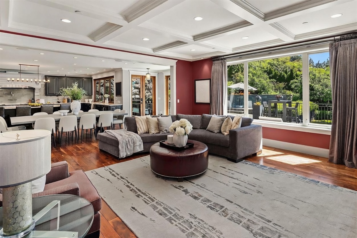 This spacious open-concept living area features a large, dark-toned sectional sofa arranged around a circular leather ottoman, set atop a textured area rug. The room is defined by a coffered ceiling and deep red accent walls, with a seamless transition into a bright dining area and kitchen beyond. Large windows provide an expansive view of the outdoor patio and lush greenery, filling the space with natural light.