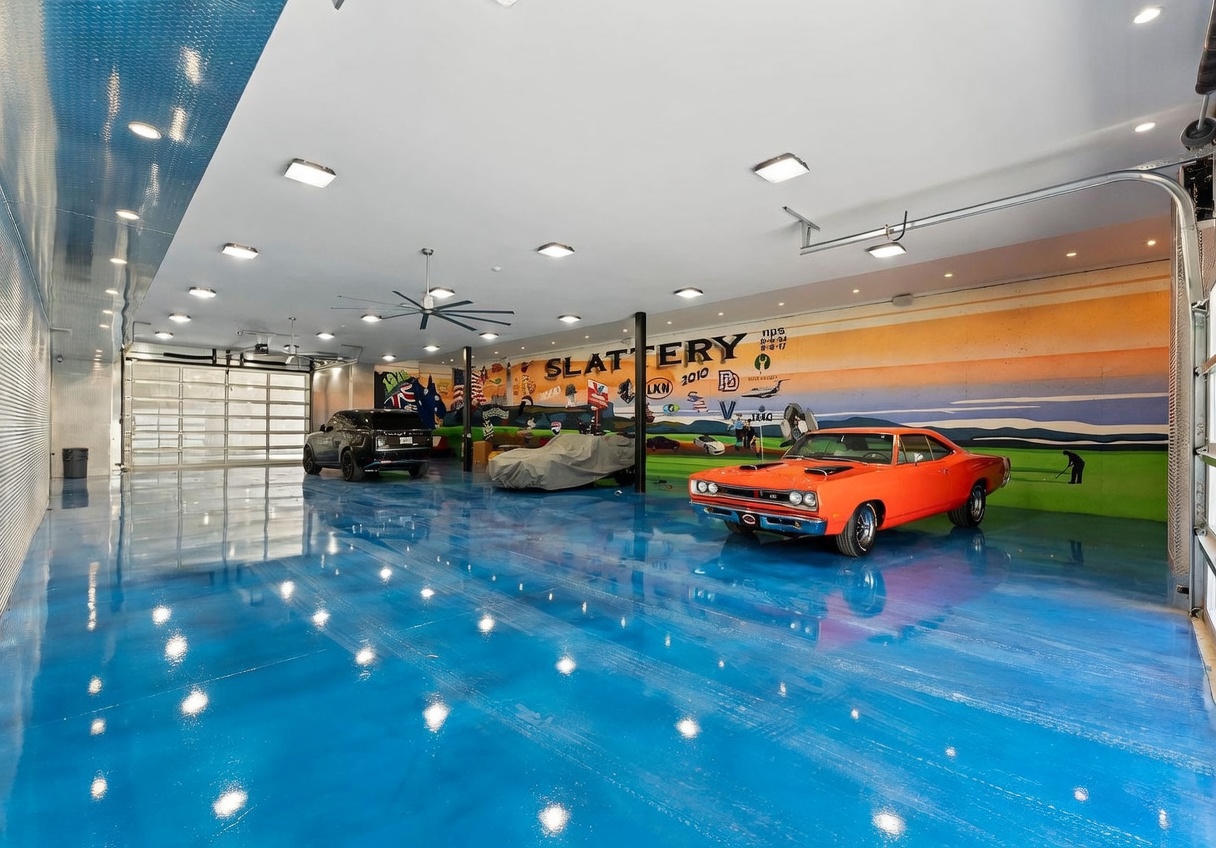 This expansive, high-ceilinged garage features a striking, high-gloss blue epoxy floor that reflects the overhead LED lighting. The space is decorated with a large, custom mural on the back wall and houses multiple vehicles, including a bright orange classic muscle car and a modern SUV. The industrial-style design is enhanced by metal-paneled walls and a large, multi-section glass garage door, creating a professional and polished automotive display area.