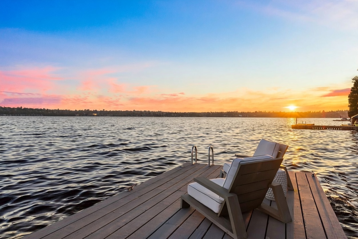 This image captures a serene, elevated perspective of a wooden dock extending into a calm lake at sunset. Two comfortable lounge chairs with white cushions are positioned on the dock, inviting relaxation while overlooking the expansive water and the distant shoreline under a vibrant, colorful sky. The scene evokes a peaceful, high-end waterfront lifestyle, emphasizing the property's direct access to the water.