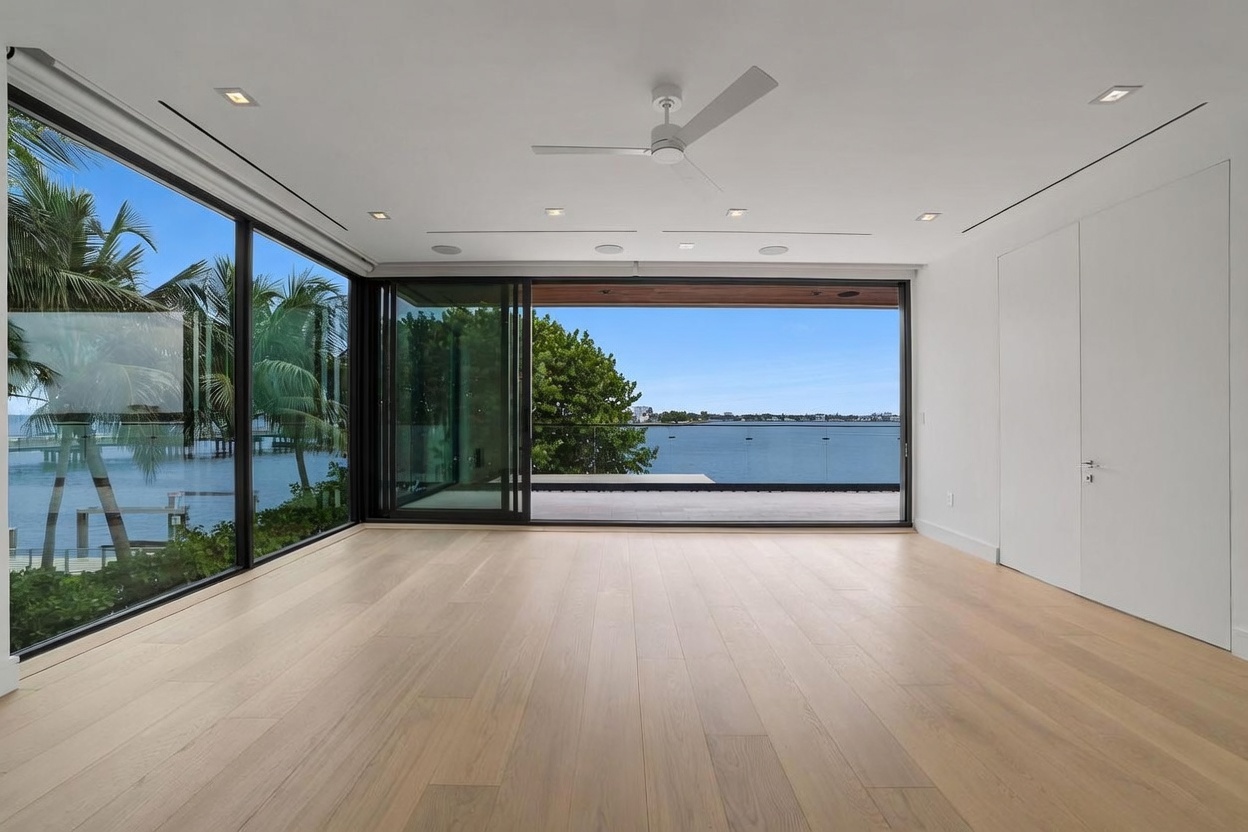 This minimalist living space features light wood flooring and crisp white walls, creating a bright and airy atmosphere. The room is defined by expansive floor-to-ceiling sliding glass doors that open to a waterfront view, seamlessly blending indoor and outdoor living. A modern ceiling fan is centered in the room, which is otherwise unfurnished, emphasizing the clean architectural lines and the scenic exterior backdrop.