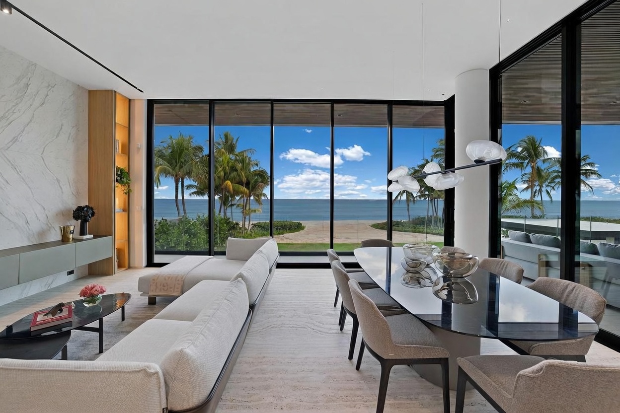 This open-concept living and dining area features a sophisticated, modern aesthetic with a neutral color palette and high-end finishes. The space is defined by expansive floor-to-ceiling glass walls that provide an unobstructed, panoramic view of the beach and ocean beyond. A sleek dining table with a dark, reflective surface sits adjacent to a comfortable seating area, creating a seamless flow perfect for entertaining while enjoying the coastal scenery.
