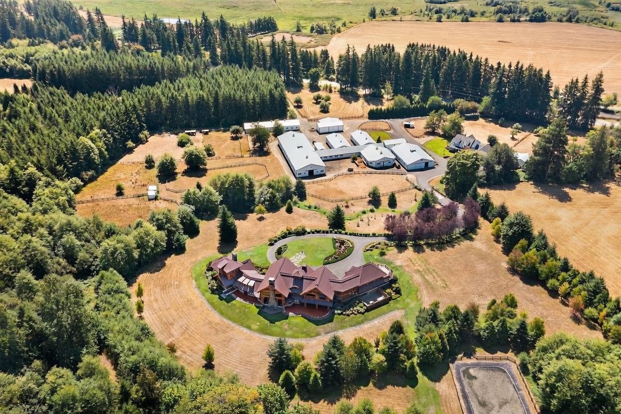 This expansive aerial view captures a sprawling rural estate featuring a large, multi-winged residence with a dark metal roof and stone accents. The property includes a circular driveway, extensive manicured lawns, and a significant complex of outbuildings and barns situated amidst dense forest and open fields. The perspective provides a comprehensive look at the vast acreage and the layout of the various structures on the grounds.