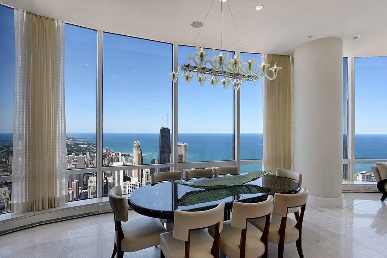This sophisticated dining room features a dark, polished oval table surrounded by six upholstered chairs, positioned beneath a distinctive, multi-armed glass chandelier. The space is defined by expansive floor-to-ceiling windows that provide a panoramic, high-altitude view of the city skyline and the lake. The room is finished with light-toned stone flooring and neutral curtains, creating an elegant and airy atmosphere suitable for formal entertaining.