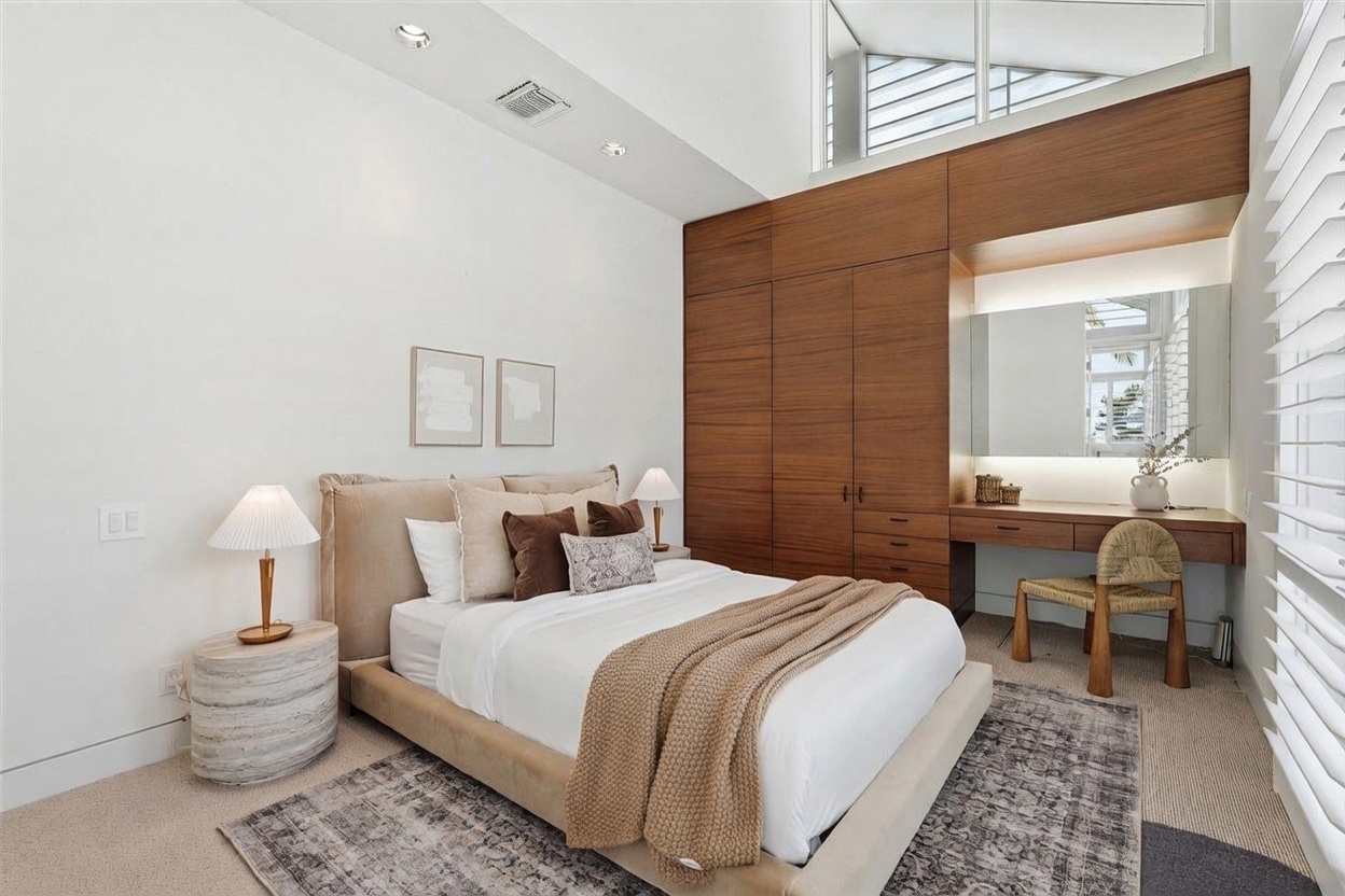 This modern bedroom features a neutral color palette with a plush upholstered bed and a textured area rug. A built-in wood cabinetry unit incorporates a vanity area with a large mirror and integrated lighting, providing a functional and stylish storage solution. The room is brightened by tall windows and high ceilings, creating an airy and sophisticated atmosphere.