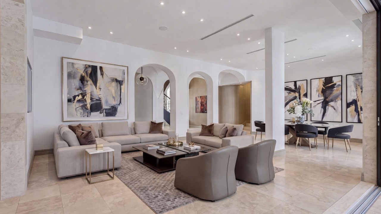 This elegant and spacious living room features a contemporary open-concept design with neutral tones, arched architectural details, and high-quality marble tile flooring. The space is anchored by a large, comfortable L-shaped sectional, complementary armchairs, and sophisticated modern artwork that ties the room together with metallic accents. The seamless transition between the living and dining areas, accented by arched transitions and recessed lighting, creates an open and luxurious atmosphere perfect for entertaining.