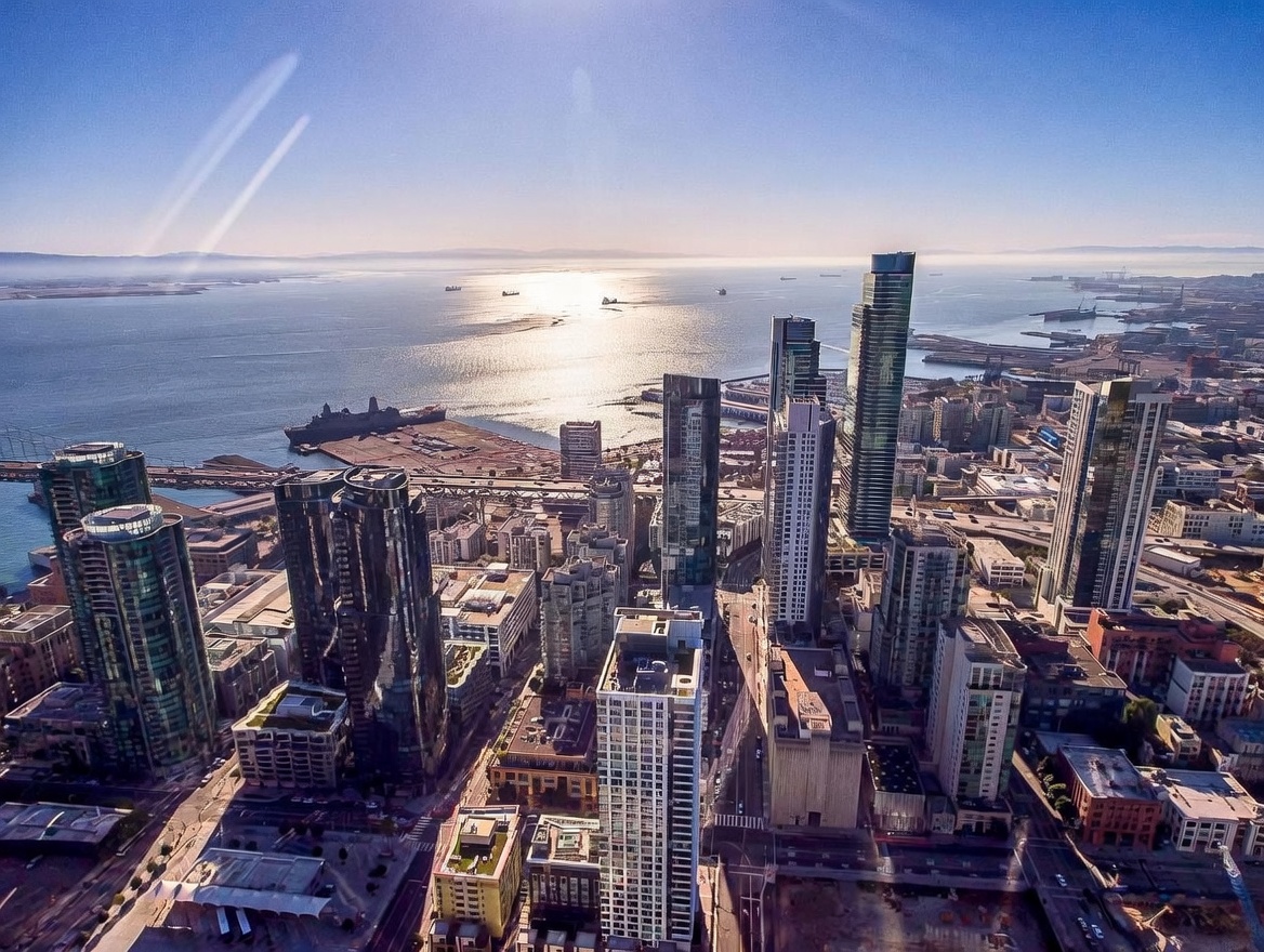 This high-altitude aerial view captures a dense urban skyline overlooking a shimmering bay under a clear blue sky. The perspective highlights a collection of modern glass skyscrapers and high-rise residential towers, with the sun reflecting brightly off the water in the distance. The composition emphasizes the scale and density of the metropolitan environment, showcasing the proximity of the city to the waterfront.