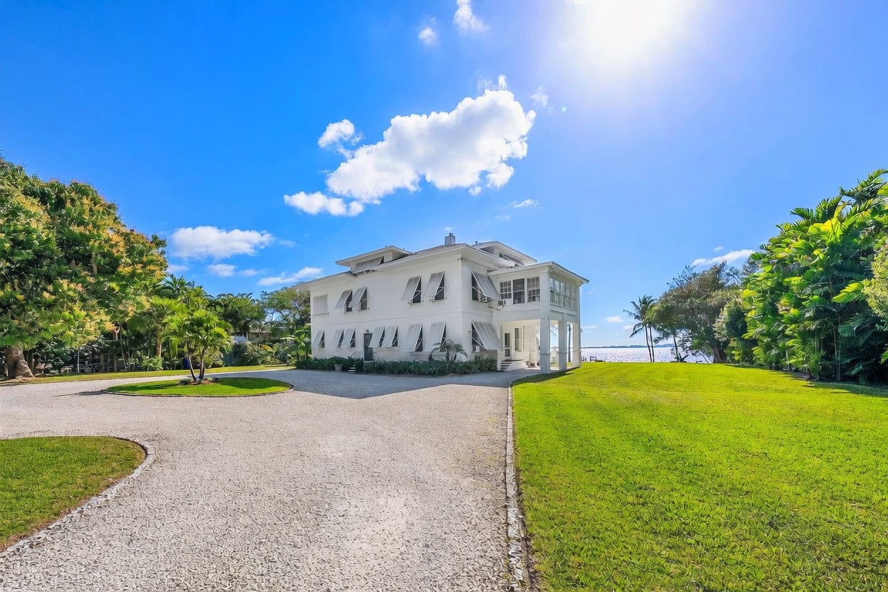 This side view captures a classic, multi-story white colonial-style home set against a vibrant blue sky with scattered clouds. The property features a sprawling gravel driveway, a manicured green lawn, and mature landscaping, with a glimpse of the water visible in the background. The architecture is defined by its symmetrical design, numerous shuttered windows, and a covered porch area, creating an elegant and serene coastal atmosphere.