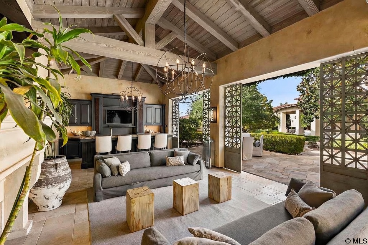 This is an interior shot of a luxurious living room that seamlessly blends indoor and outdoor living. The room features a high, beamed ceiling, a gray sofa, and wooden block tables, creating a rustic yet elegant atmosphere. Large, ornate metal doors open to a beautifully landscaped patio and garden, enhancing the sense of spaciousness and connection to nature.