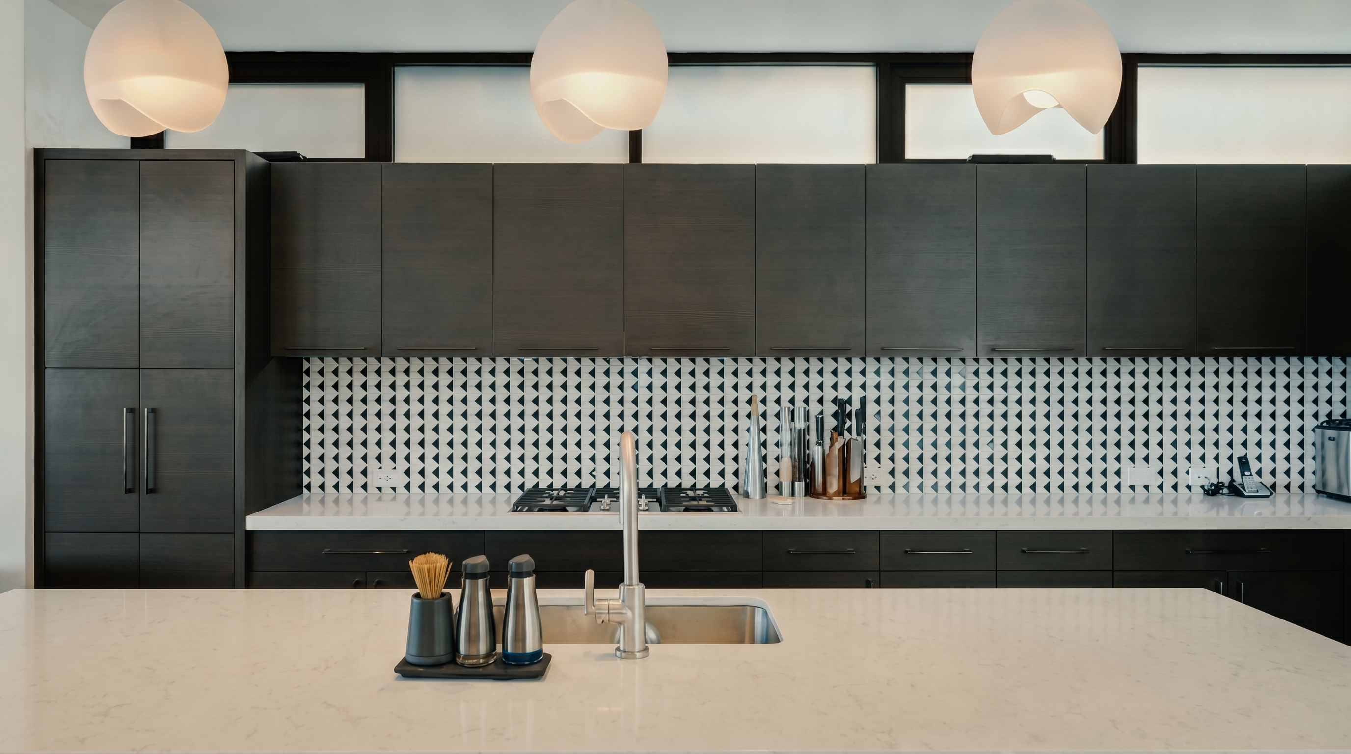 This modern kitchen features dark, flat-panel cabinetry paired with a striking geometric patterned tile backsplash. A large central island with a light-toned countertop and an integrated sink serves as the focal point, illuminated by three contemporary pendant lights. The space is framed by horizontal clerestory windows that allow natural light to filter in above the upper cabinets.