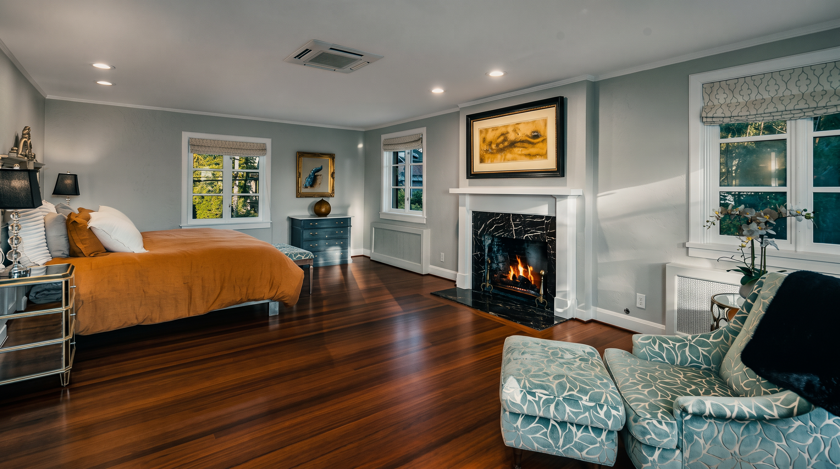 This spacious primary bedroom features rich, polished wood flooring and light-toned walls, creating a warm and inviting atmosphere. A prominent fireplace with a dark stone surround and a white mantel serves as a focal point, complemented by a comfortable seating area with a patterned armchair and ottoman. The room is well-lit by recessed lighting and large windows that provide ample natural light, while a plush bed with an orange duvet adds a vibrant pop of color to the space.