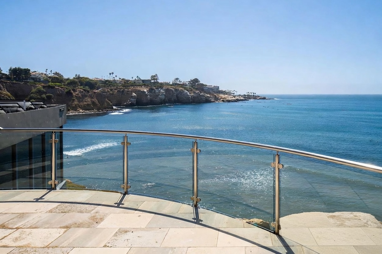This image captures a sweeping, curved balcony featuring stone tile flooring and a modern glass-paneled railing with stainless steel posts. The vantage point offers a panoramic, cinematic view of a rugged coastline, deep blue ocean, and distant cliffs under a clear sky. The perspective emphasizes the seamless transition between the private outdoor living space and the expansive natural scenery.