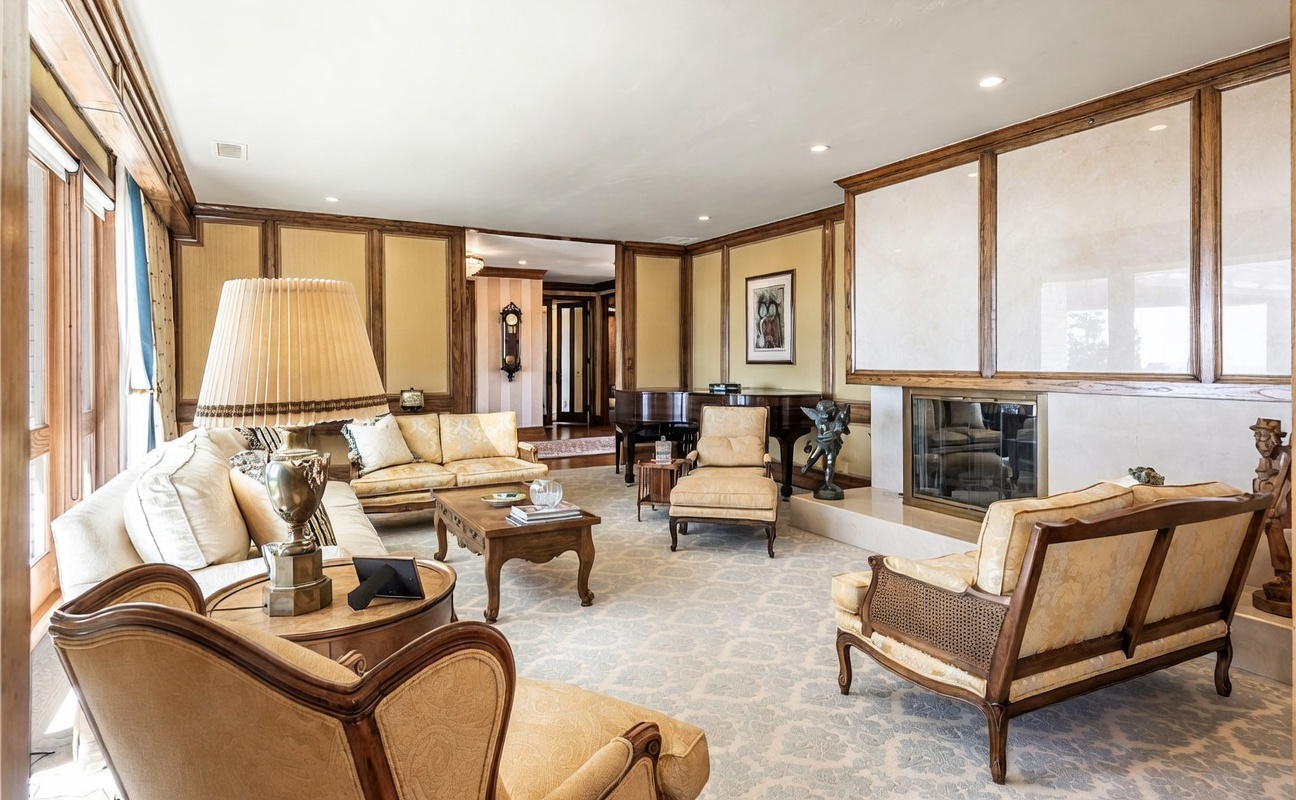This elegant, traditional living room features wood-paneled walls and a neutral patterned carpet. The space is furnished with multiple upholstered sofas and armchairs in a coordinated light fabric, centered around a wooden coffee table and a grand piano in the background. A large fireplace with a prominent surround serves as a focal point, while the room is illuminated by a combination of recessed lighting and a decorative table lamp.