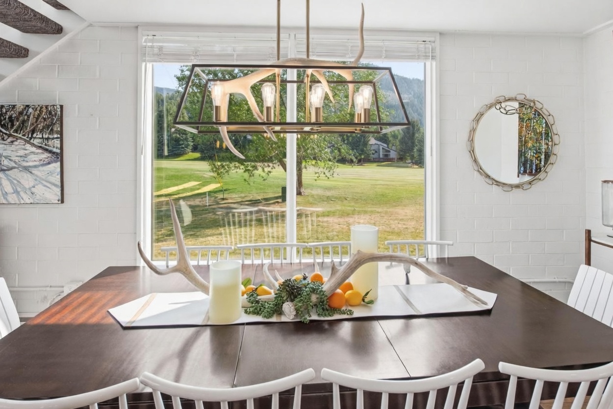 This dining room features a dark wood table centered beneath a unique antler-inspired chandelier, creating a rustic yet refined atmosphere. A large window provides a clear view of the green landscape outside, while white painted brick walls and a circular decorative mirror add texture and brightness to the space. The table is set with a centerpiece of antlers, greenery, and citrus fruits, emphasizing a natural and inviting aesthetic.