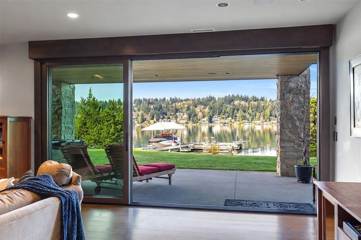 This interior living space features a seamless transition to the outdoors through expansive, floor-to-ceiling sliding glass doors that frame a serene lake view. The room is anchored by a comfortable sofa in the foreground, while the covered patio outside holds lounge chairs overlooking a private dock and boat lift. The combination of warm wood flooring and natural stone accents creates a sophisticated, tranquil atmosphere perfect for waterfront living.