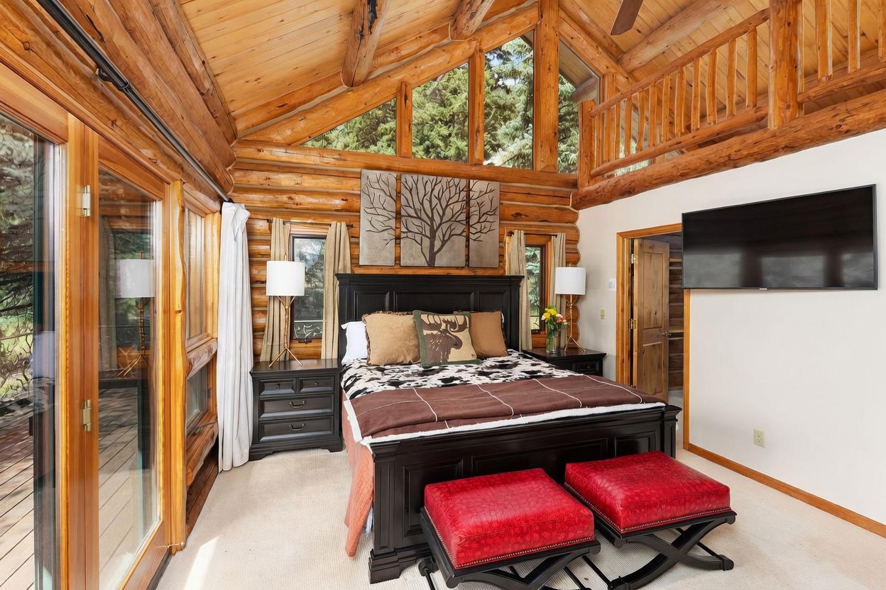 This rustic primary bedroom features exposed log walls and a vaulted ceiling with exposed timber beams, creating a warm, cabin-like atmosphere. A dark wood bed frame is centered against the wall, flanked by matching nightstands and topped with a patterned quilt, while two vibrant red ottomans sit at the foot of the bed. Large windows and a glass door provide ample natural light and views of the surrounding trees, complemented by a lofted balcony area overlooking the space.