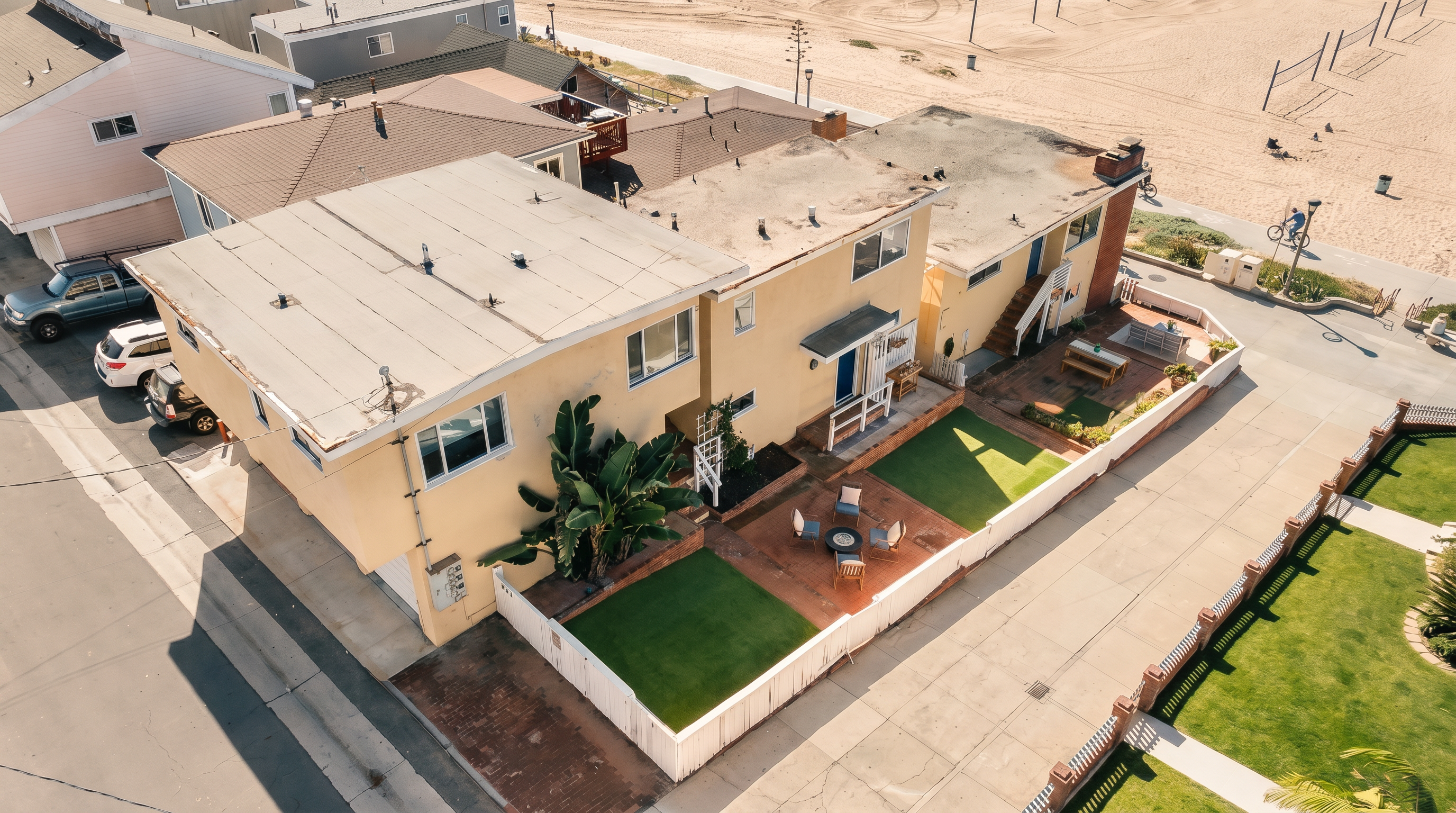 This high-angle aerial shot captures a multi-level residential property situated directly adjacent to a sandy beach. The home features a light-colored stucco exterior, a flat roof, and a private courtyard area with a patio and artificial turf. The perspective highlights the home's prime coastal location, showing the proximity to the beach path and the surrounding neighborhood layout.