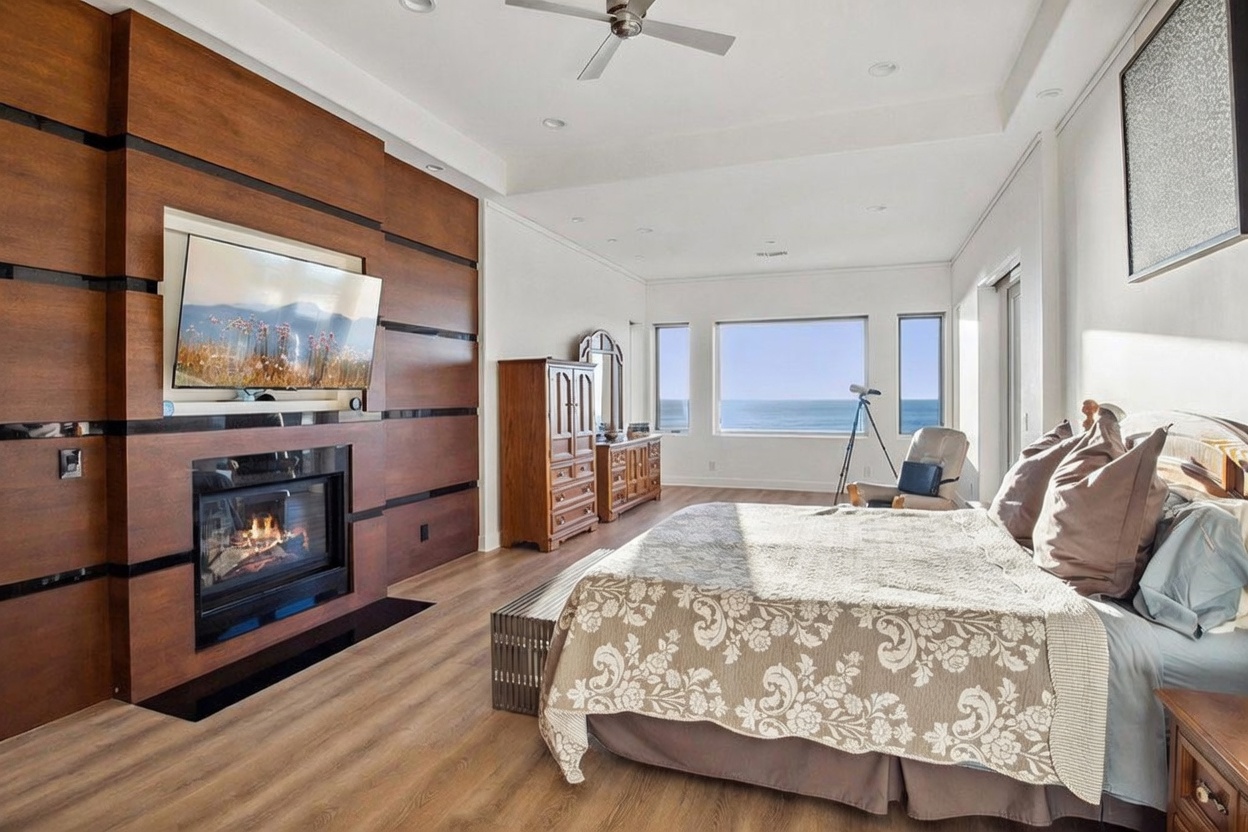 This spacious primary bedroom features a prominent wood-paneled accent wall housing a television and a fireplace with a dark surround. A large bed with patterned bedding sits in the foreground, while expansive windows offer a clear, panoramic view of the ocean. The room is finished with light wood-toned flooring and a ceiling fan, creating a serene and luxurious coastal atmosphere.