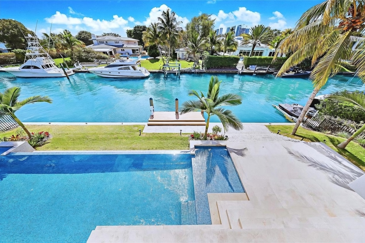 This elevated perspective captures a luxurious waterfront property featuring a rectangular swimming pool with a sun shelf overlooking a private canal. The scene includes a manicured lawn, a wooden boat dock, and several yachts moored in the turquoise water, framed by lush palm trees. The bright, sunny atmosphere highlights the high-end coastal lifestyle and the seamless transition between the outdoor living space and the waterway.