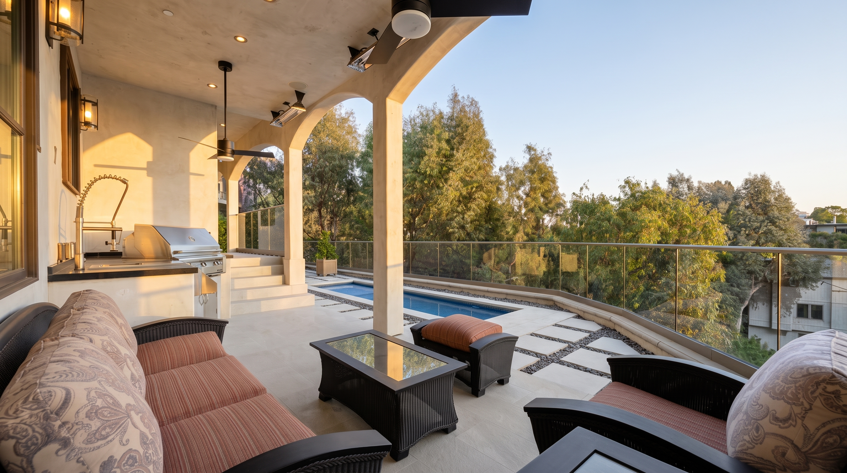 This expansive covered balcony offers a sophisticated outdoor living space complete with a built-in kitchen area, including a stainless steel grill and sink. The area is furnished with comfortable wicker-style seating and a glass-topped coffee table, overlooking a sleek lap pool and lush greenery beyond a modern glass railing. The perspective captures the seamless transition between the shaded lounge area and the sun-drenched pool deck, emphasizing a luxurious, resort-like atmosphere.