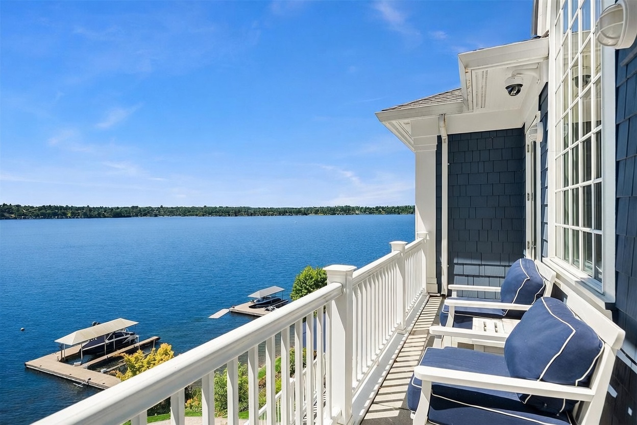 This image captures a serene, elevated balcony view overlooking a expansive, calm lake on a bright, sunny day. The balcony features white railings and comfortable outdoor seating with blue cushions, creating a perfect spot for relaxation. Below, private boat docks extend into the water, emphasizing the waterfront lifestyle and peaceful atmosphere of the property.