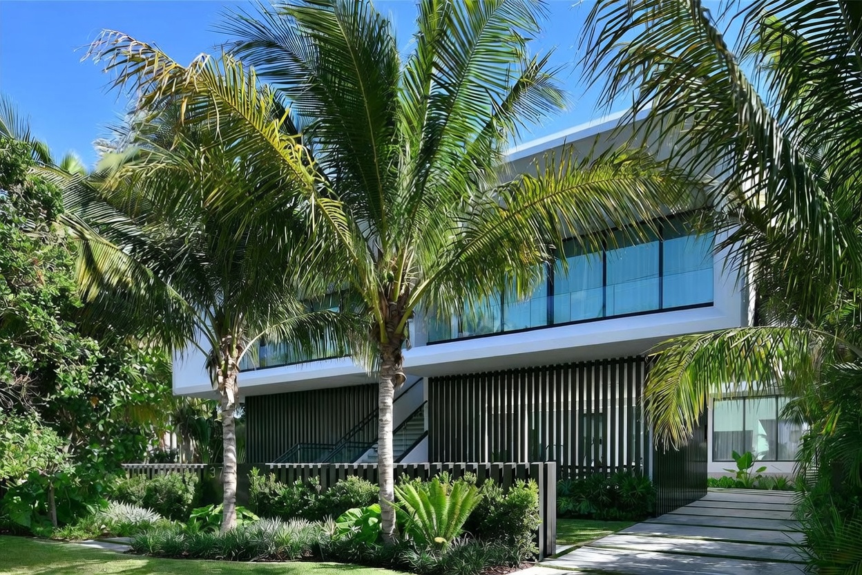 This modern two-story residence features a striking architectural design with clean lines and a prominent upper level encased in floor-to-ceiling glass. The ground floor is accented by vertical dark slats, creating a sophisticated contrast against the white exterior structure. Lush tropical landscaping, including mature palm trees, frames the property, while a paved walkway leads toward the entrance, emphasizing a contemporary and luxurious aesthetic.