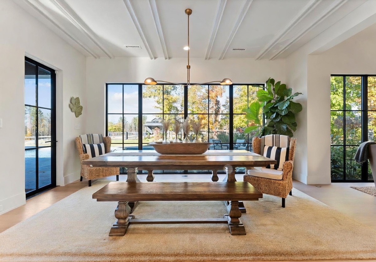 This elegant dining room features a large wooden table paired with a matching bench and two woven armchairs with striped cushions. The space is anchored by a neutral area rug and illuminated by a modern, multi-arm chandelier suspended from a coffered ceiling. Large windows with black frames provide an expansive view of the outdoor landscape, creating a bright and airy atmosphere.