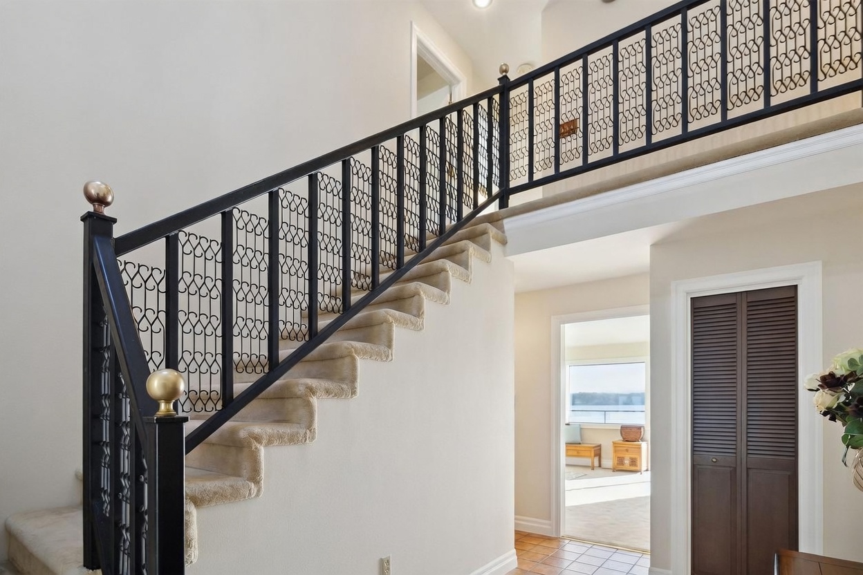 This interior view captures a grand staircase featuring a black metal railing with decorative scrollwork and gold-toned finials. The stairs are carpeted in a neutral tone, leading up to a landing that overlooks the entryway. To the right, a dark-toned louvered door provides access to a closet, while an open doorway in the background reveals a glimpse of a bright room with a view of the outdoors.