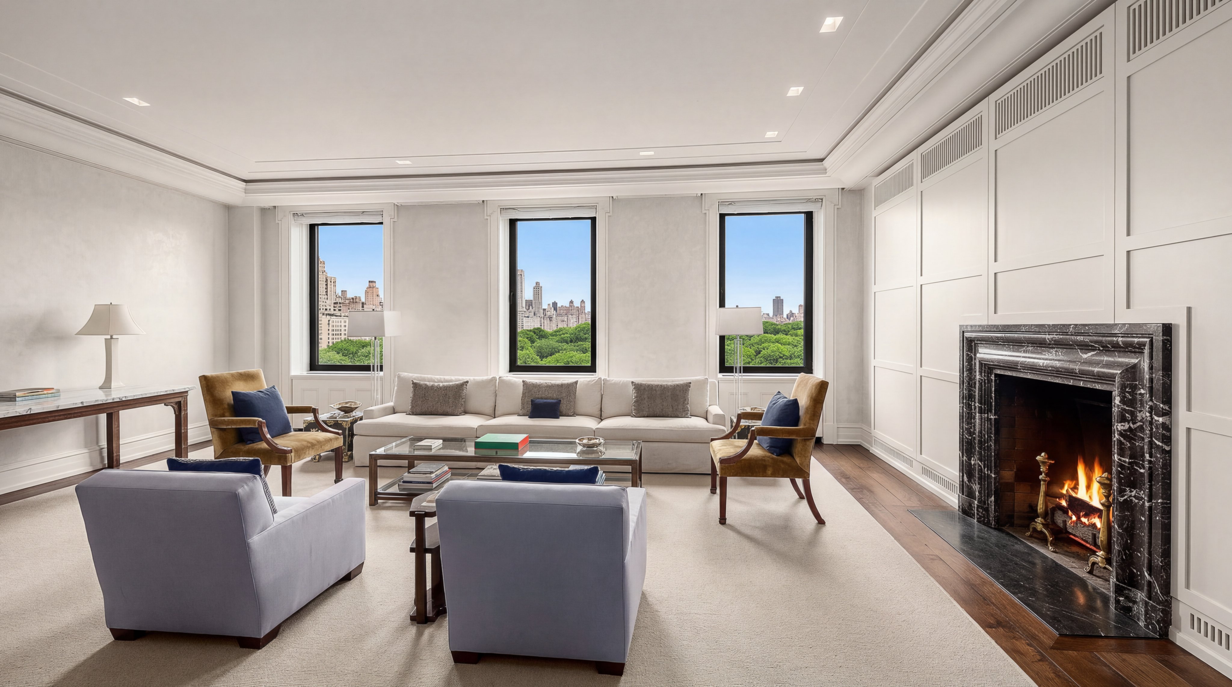 This elegant and spacious living room features a classic aesthetic with a white paneled fireplace mantel as its centerpiece, housing a cozy, lit fire. The room is light-filled, thanks to three large windows framing a scenic view of lush treetops and the city skyline. Meticulously arranged neutral-toned plush seating, complemented by soft blue accent chairs and a central glass-topped coffee table, creates a sophisticated atmosphere perfect for luxury urban living.