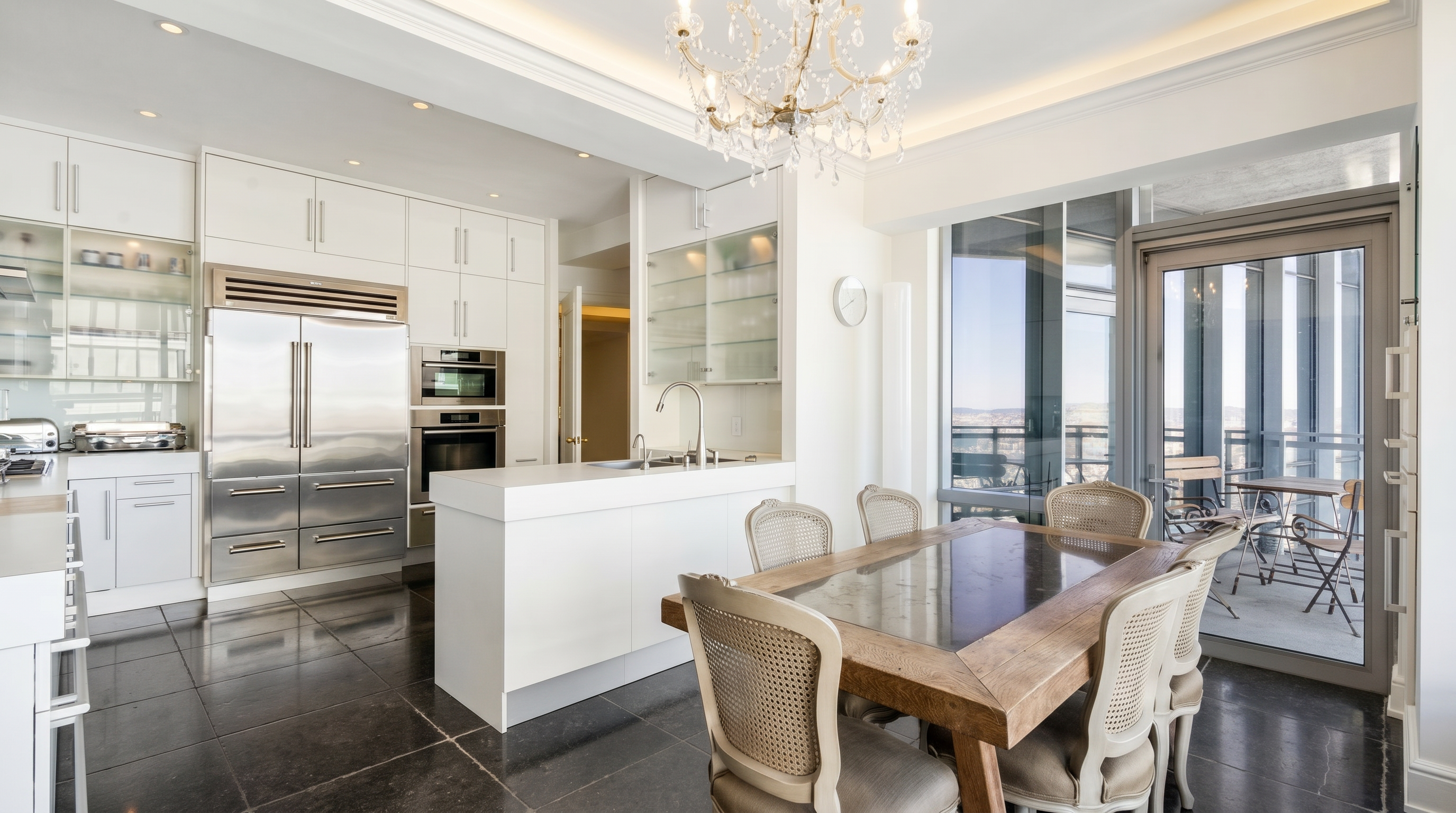 This bright, open-concept kitchen and dining area features a sleek white island and cabinetry paired with dark stone flooring. A rustic wooden dining table with upholstered chairs sits adjacent to large windows that provide natural light and access to an outdoor balcony. The space is anchored by a crystal chandelier, creating a sophisticated blend of modern and traditional design elements.